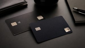 A imagem complementa esse cenário ao apresentar uma composição minimalista e elegante com dois cartões escuros posicionados sobre uma mesa sofisticada. Elementos como caderno, caneta e acessórios executivos reforçam a ideia de exclusividade, alto padrão e ambiente financeiro premium. A estética limpa, moderna e sem excesso de informações visuais transmite sofisticação e representa visualmente a concorrência entre instituições que disputam consumidores de elevado poder aquisitivo.