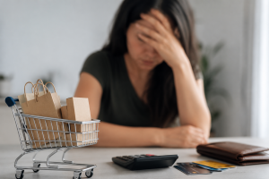 Mulher sentada à mesa com expressão de preocupação, apoiando a mão na testa enquanto observa itens relacionados às finanças pessoais. Em primeiro plano, um carrinho de compras em miniatura com sacolas de papel aparece ao lado de cartões de crédito, carteira e calculadora sobre uma superfície clara. O ambiente interno possui iluminação suave, tons neutros e composição minimalista, transmitindo a sensação de sobrecarga financeira, consumo impulsivo e dificuldades no controle do orçamento doméstico. A cena ilustra os impactos emocionais do parcelamento e da gestão das finanças no contexto brasileiro.